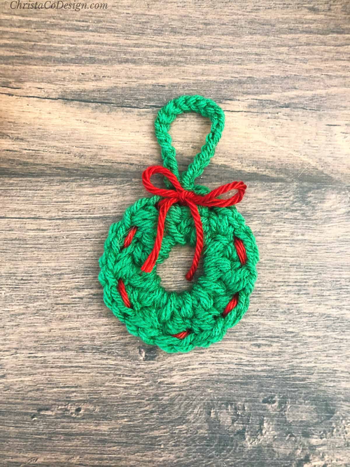 Green crochet wreath ornament with red bow on wood table.