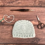 Light blue crochet baby hat on wood table with notions and heart.