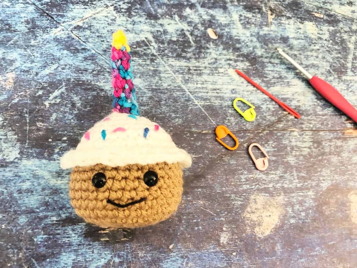 Crochet cupcake on blue background with stitch markers, needle and hook.