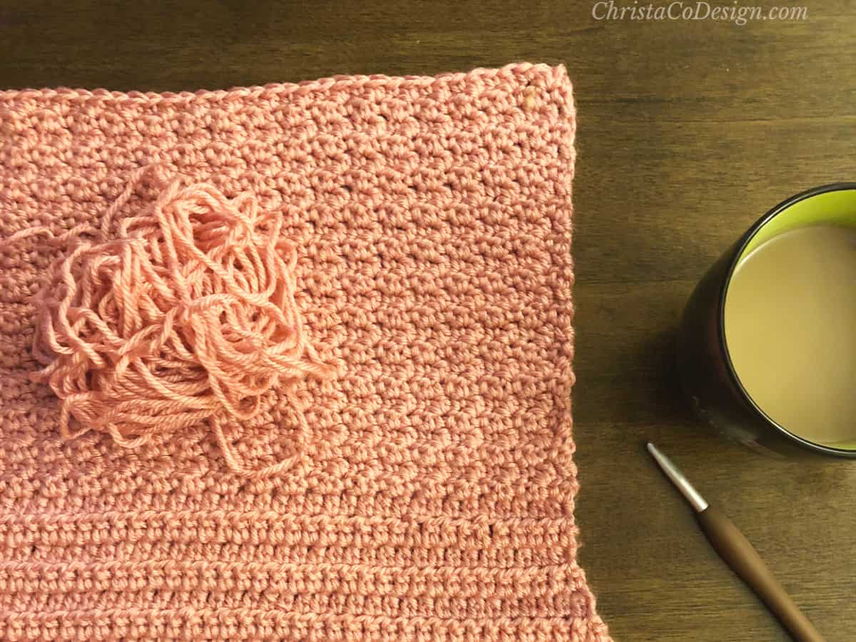 Pink crochet blanket with uneven edge on table with hook and coffee.