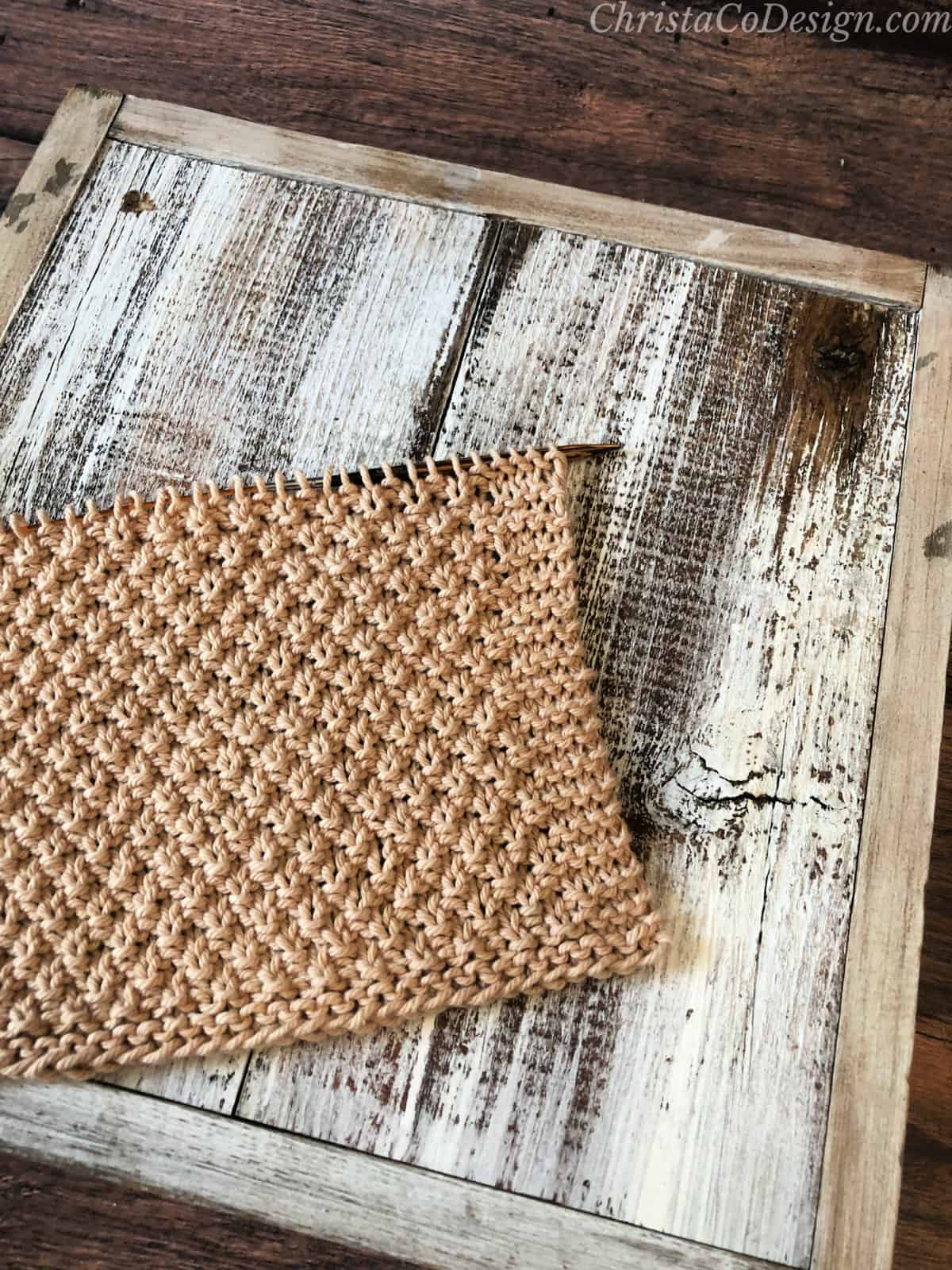 Brown textured dishcloth on the knitting needles on white wood box. 