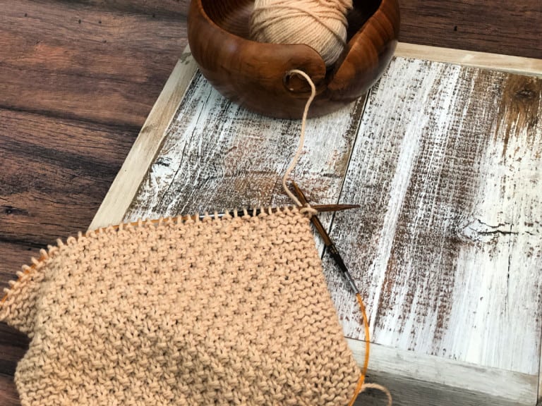 Brown dishcloth on knitting needles on white wood.