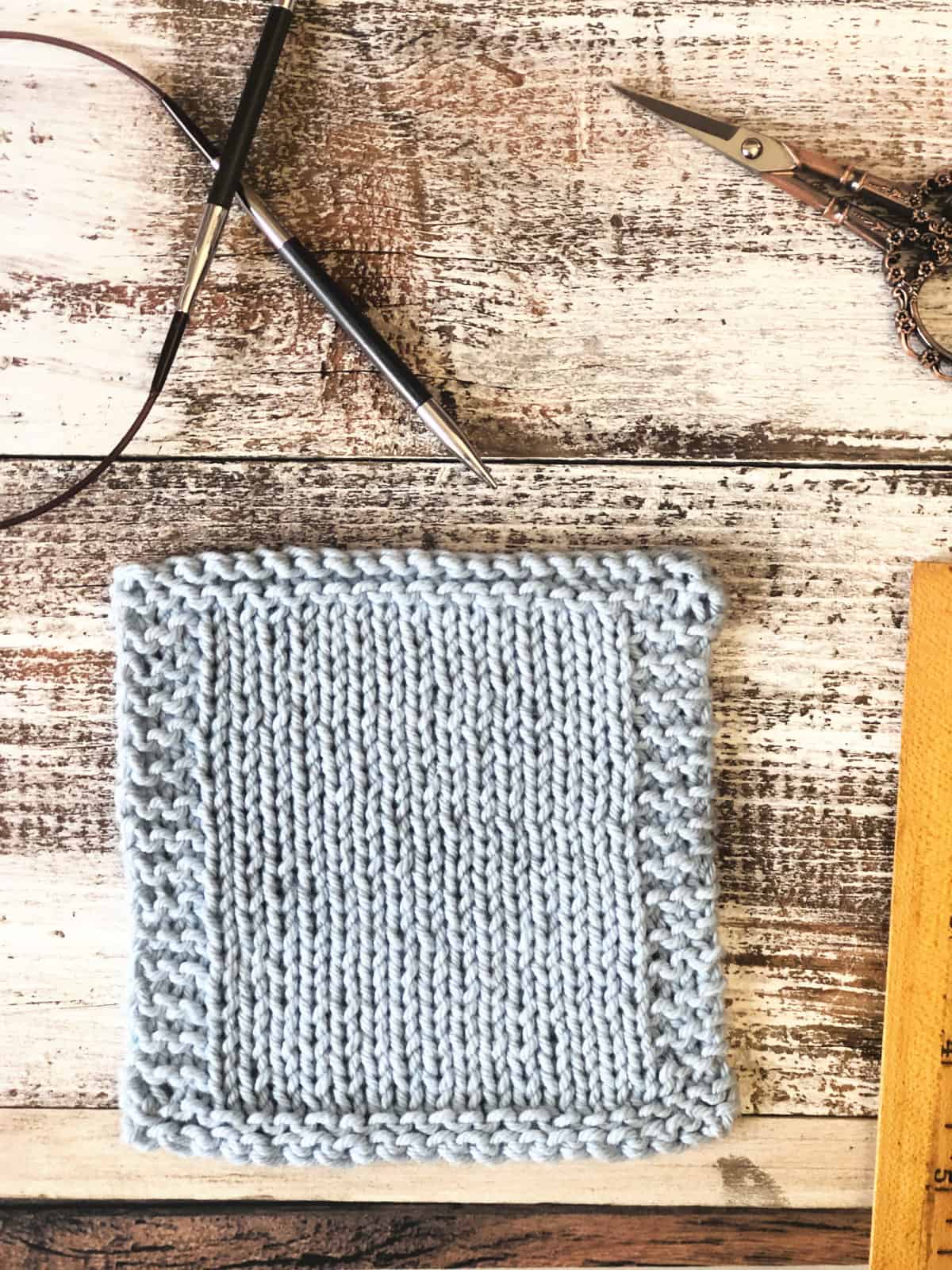 Knitting needle, scissors, ruler next to blue knit coaster.
