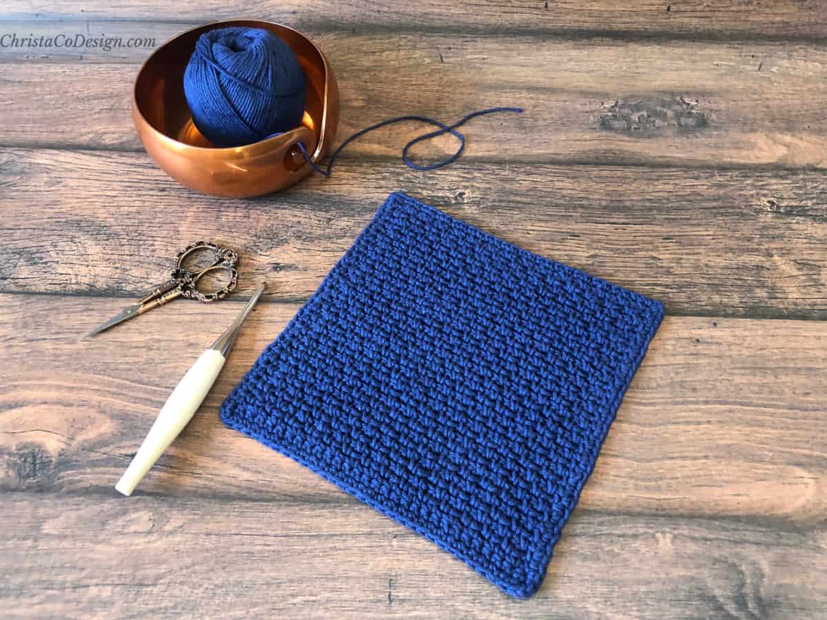 Blue square dishcloth with crochet hook, scissors and yarn bowl.