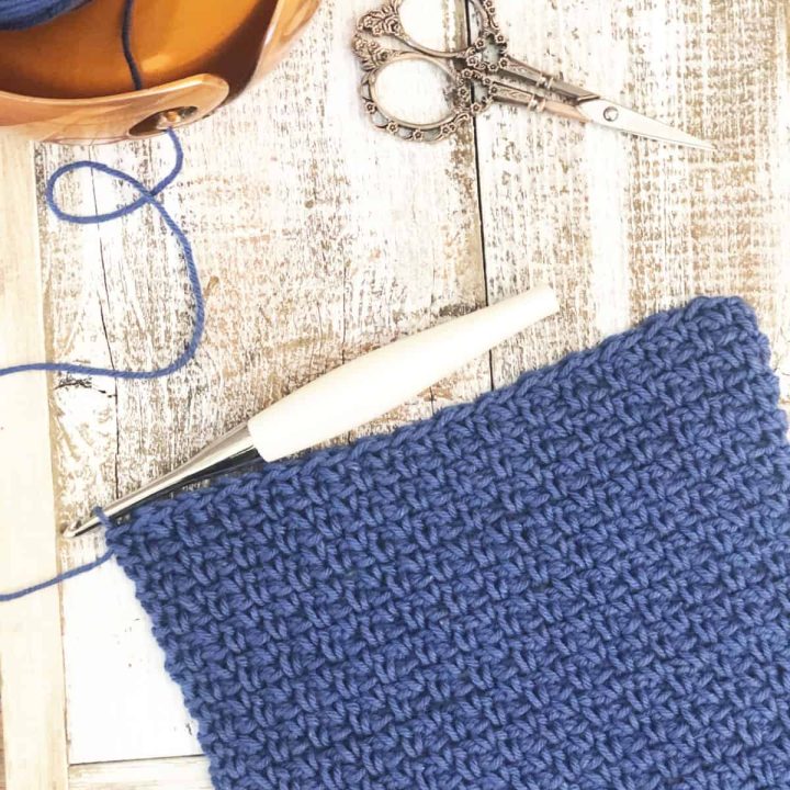 Blue crochet square in moss stitch with hook, scissors and yarn bowl.