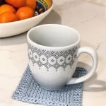 White coffee cup with geometric design in grey on pale blue knit coaster next to bowl or clementines.