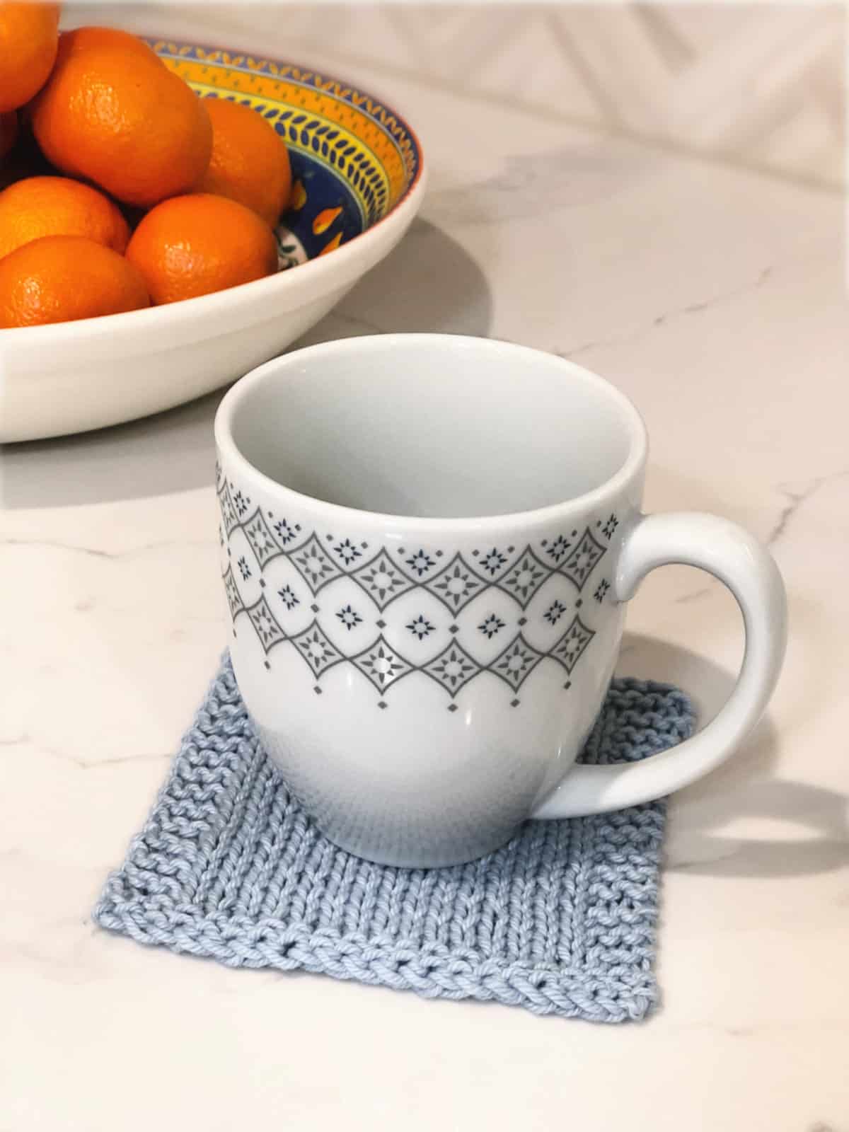 White coffee cup with geometric design in grey on pale blue knit coaster next to bowl or clementines.