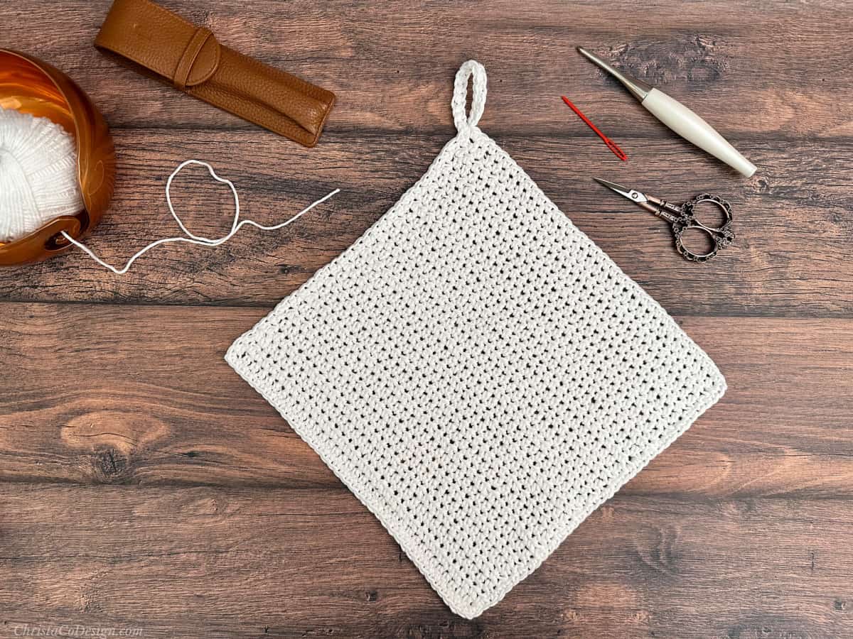 White crochet dishcloth with loop, hook, scissors and yarn bowl.
