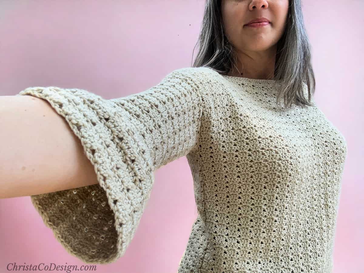 Woman in crochet sweater with arm extended showing bell sleeves.