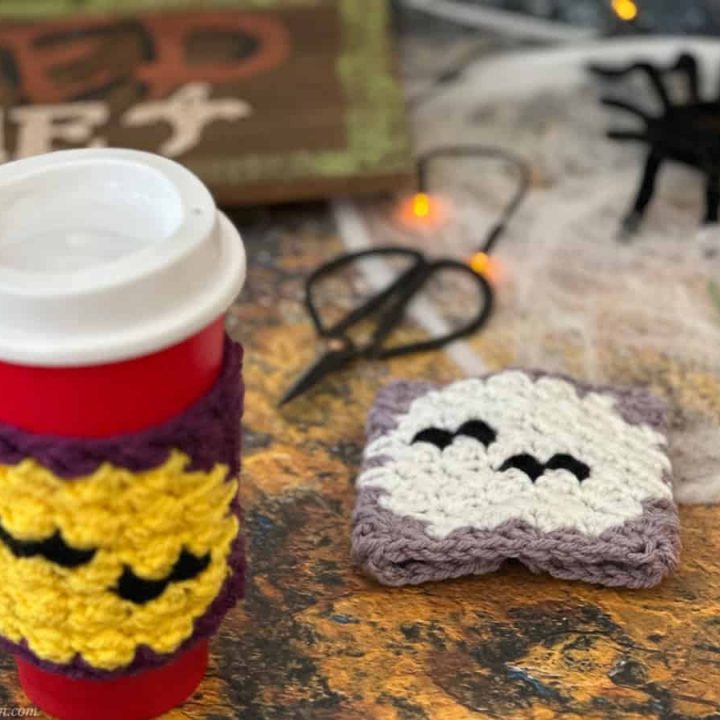 Crochet cup cozies in purple with bats flying over moon among halloween decorations.