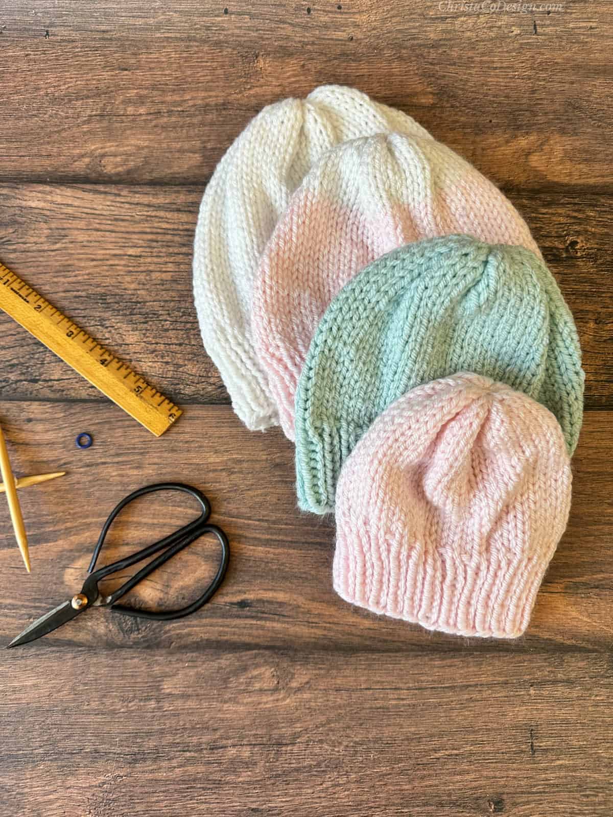 Four baby hats in white, pink and blue stacked with knitting notions.