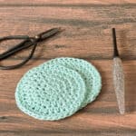 Round crochet scrubbiest in mint yarn next to glitter hook and scissors.