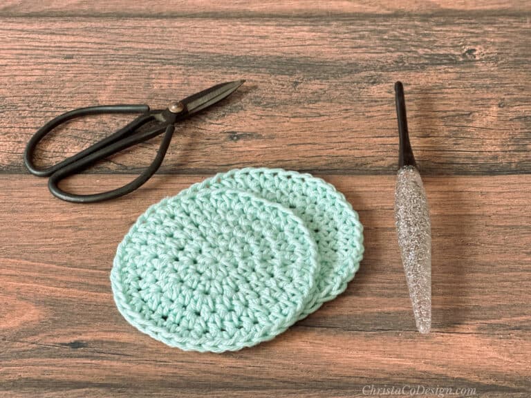 Round crochet scrubbiest in mint yarn next to glitter hook and scissors.