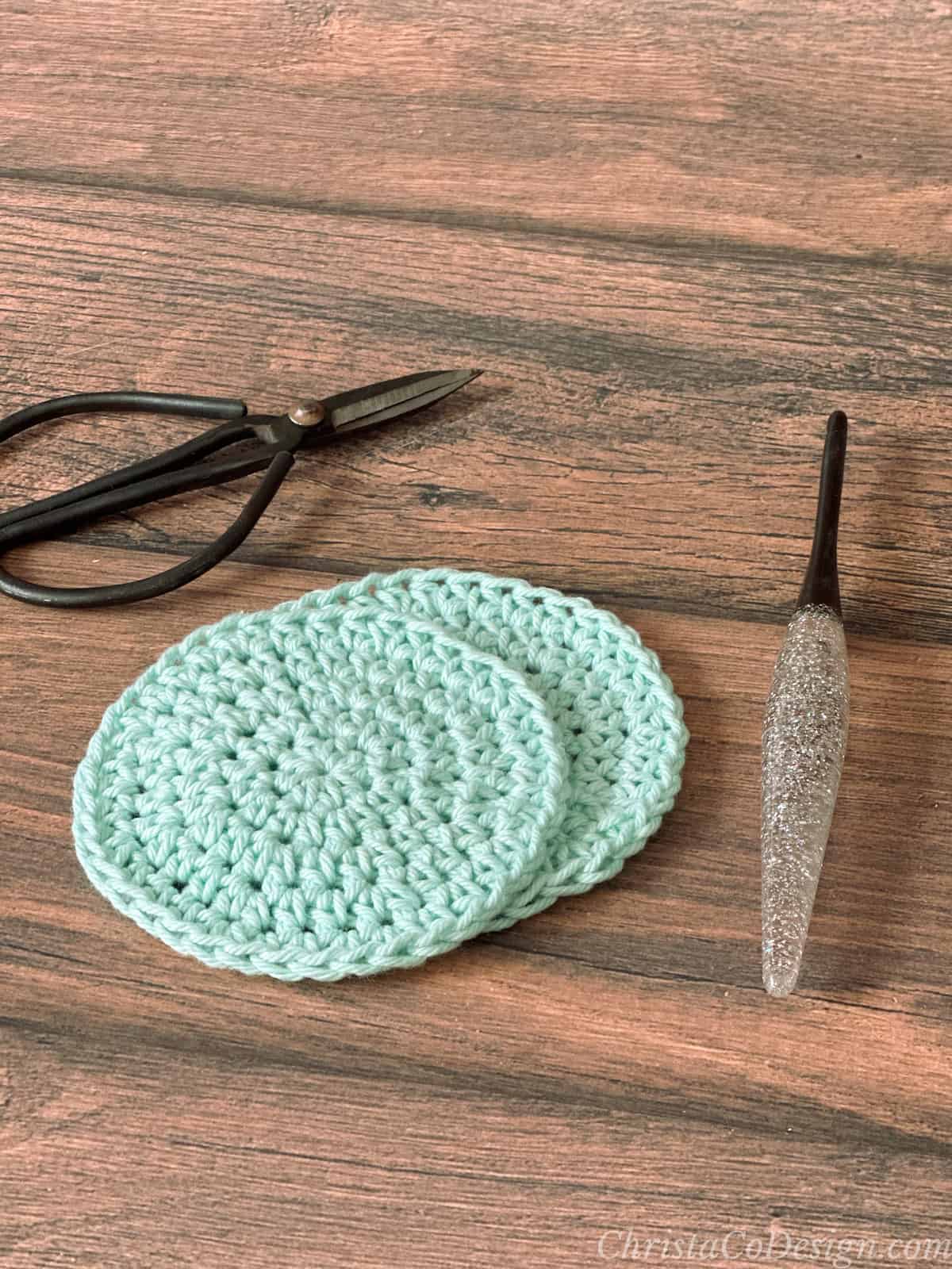 Two mint colored round crochet scrubbiest next to scissors and crochet hook.