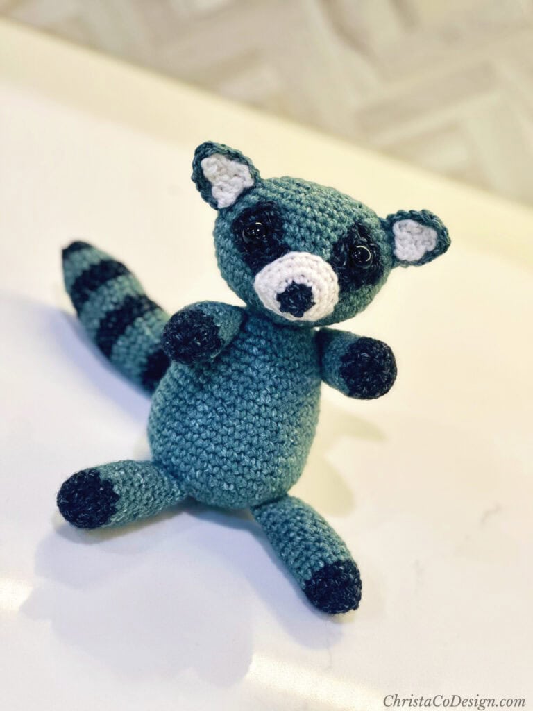 Crochet raccoon amigurumi sitting on white table.