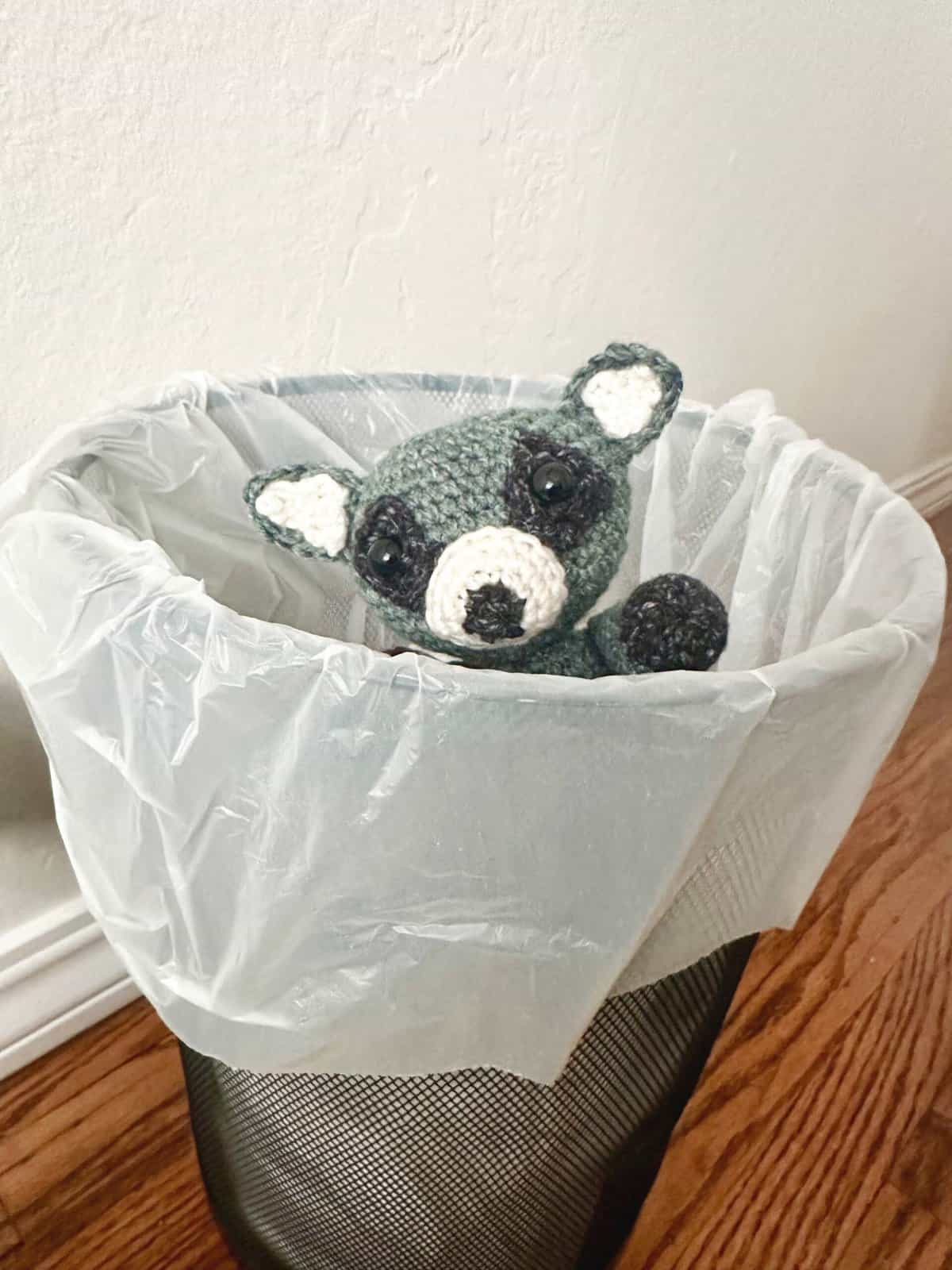 Crochet raccoon peeking out of trashcan.