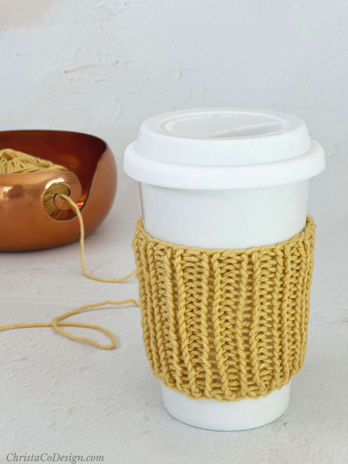 White coffee cup with yellow knit ribbed cozy and yarn in bowl.