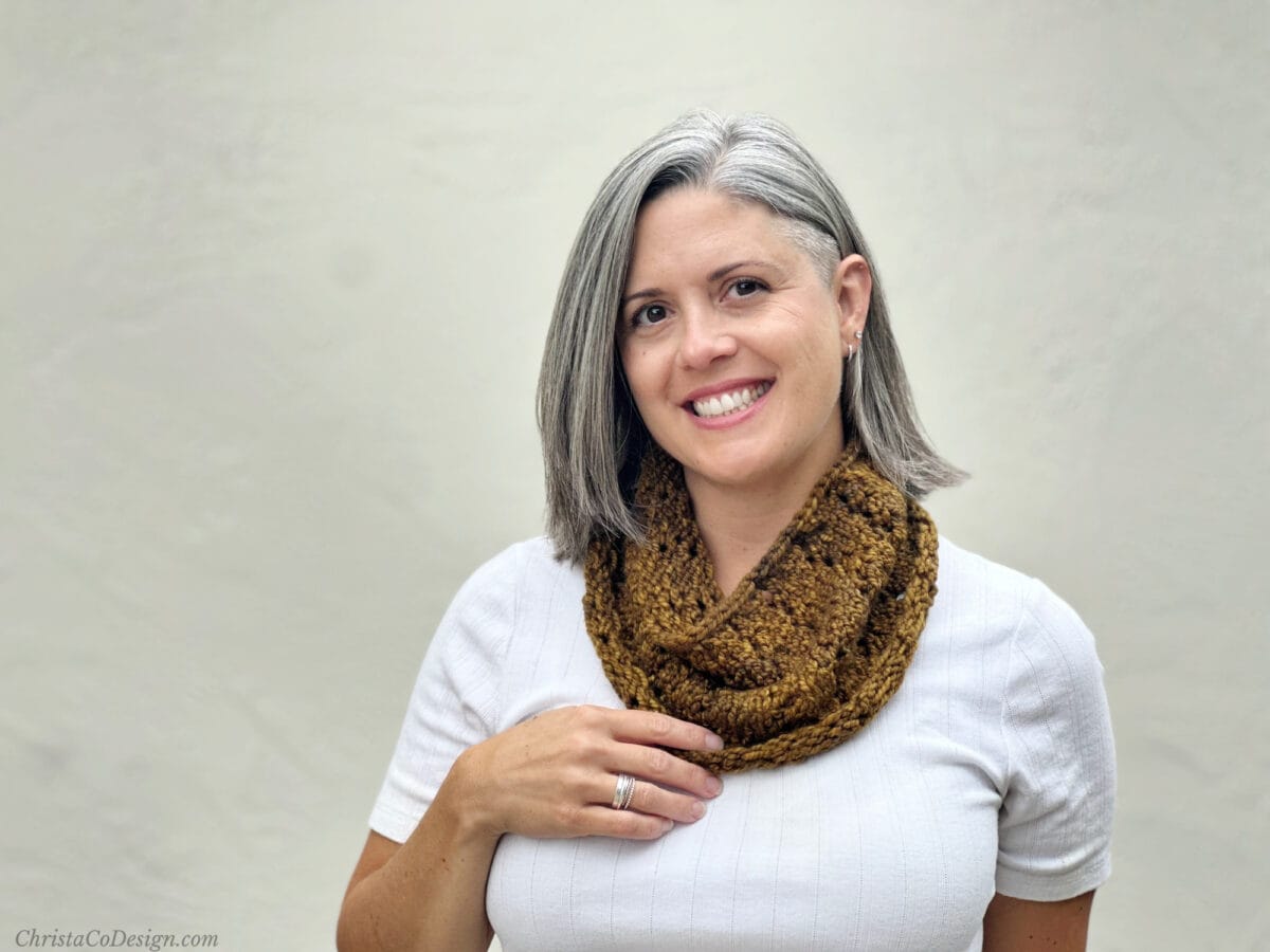 Woman in white top smiling with hand on knit cowl in dark mustard.