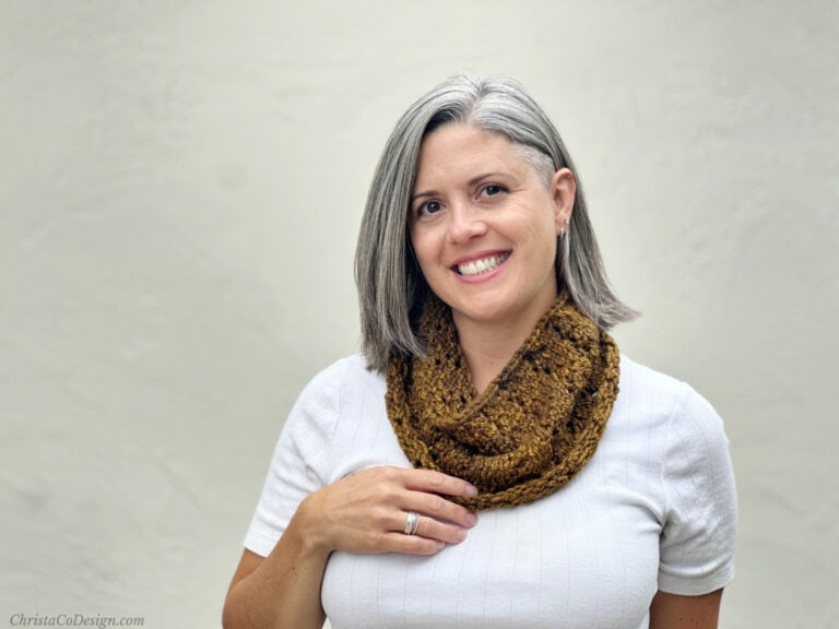 Woman in white top smiling with hand on knit cowl in dark mustard.