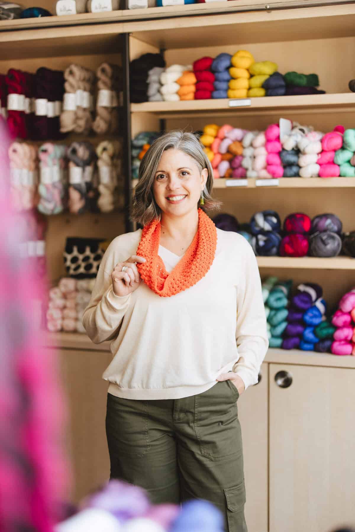 Woman smiling with coral colored knit cowl in yarn shop.