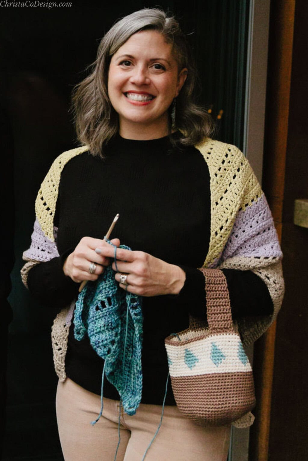 Woman smiling with crochet bag on arm with hook and shawl.