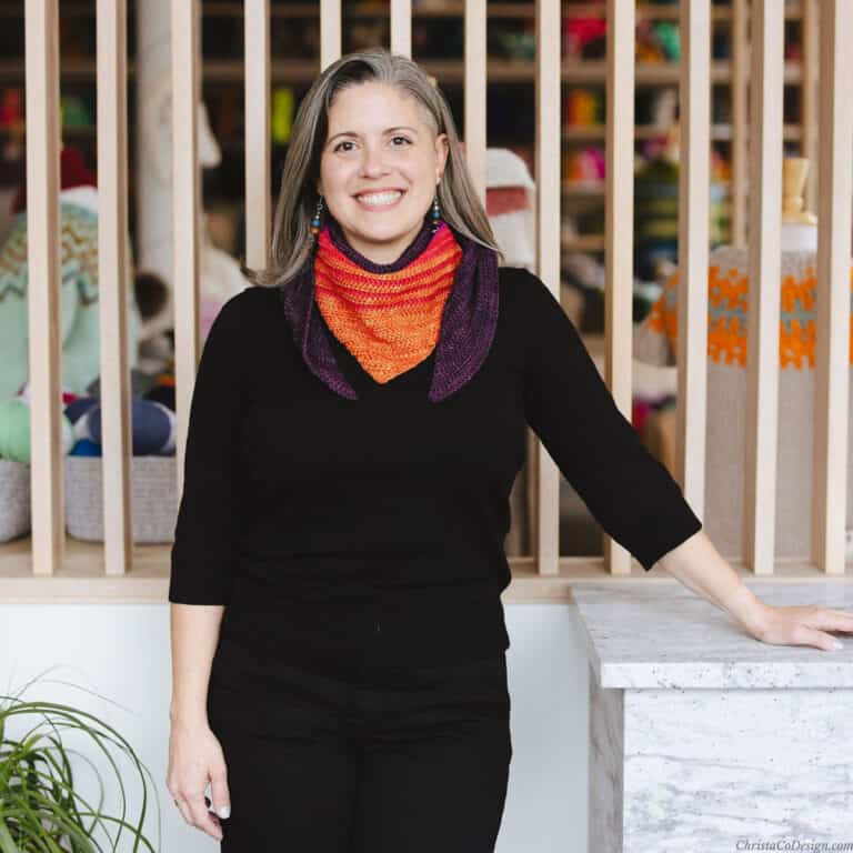 Woman leaning on marble counter with crochet scarf in oranges.