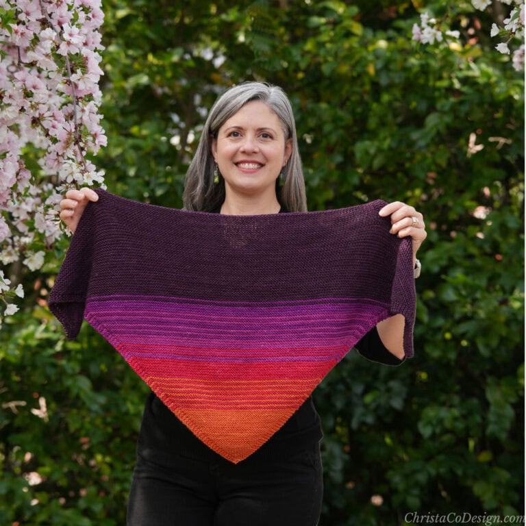 Woman smiling holding open knit triangle scarf in colors of the sunset.