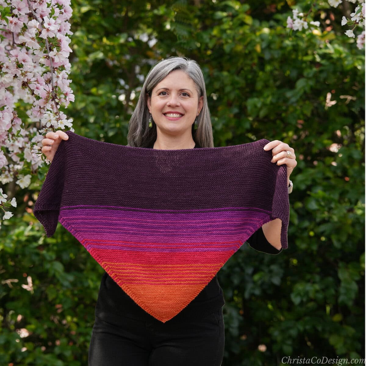 Woman smiling holding open knit triangle scarf in colors of the sunset.