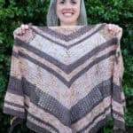 Smiling woman holding crochet shawl in pink and brown stripes.