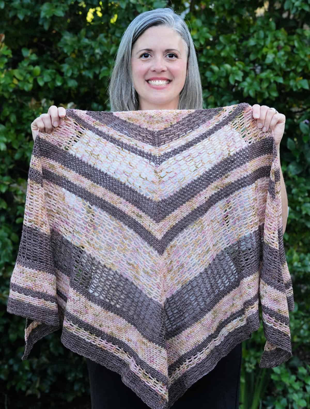 Smiling woman holding crochet shawl in pink and brown stripes.