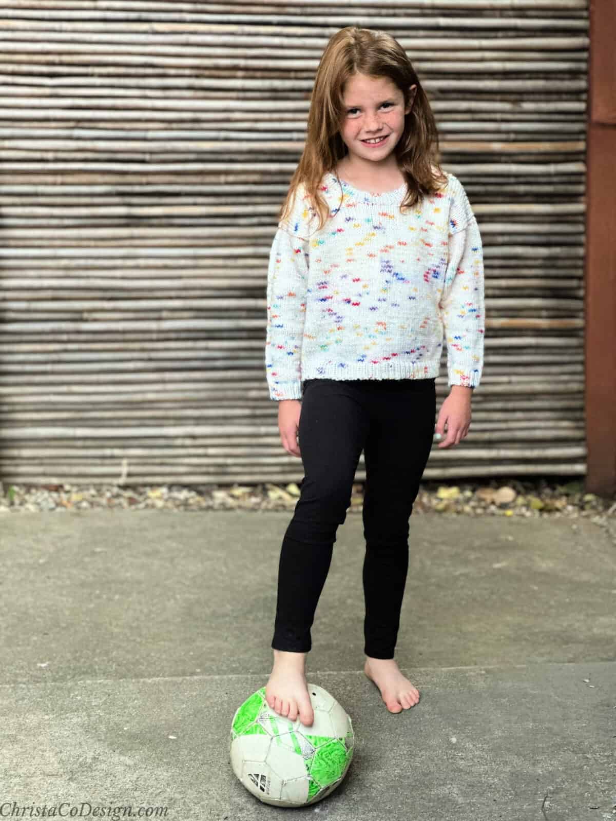 Girl in colorful knit sweater with foot on soccer ball.