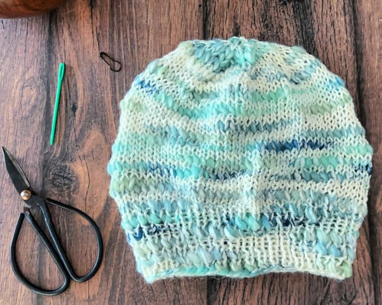 Flay lay of knit hat in blues, greens and white next to knitting notions.